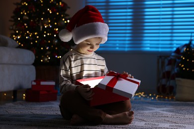 Photo of Little boy in Santa hat with Christmas gift on floor at home