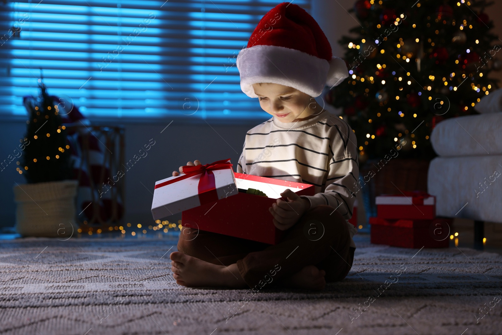 Photo of Little boy in Santa hat with Christmas gift on floor at home