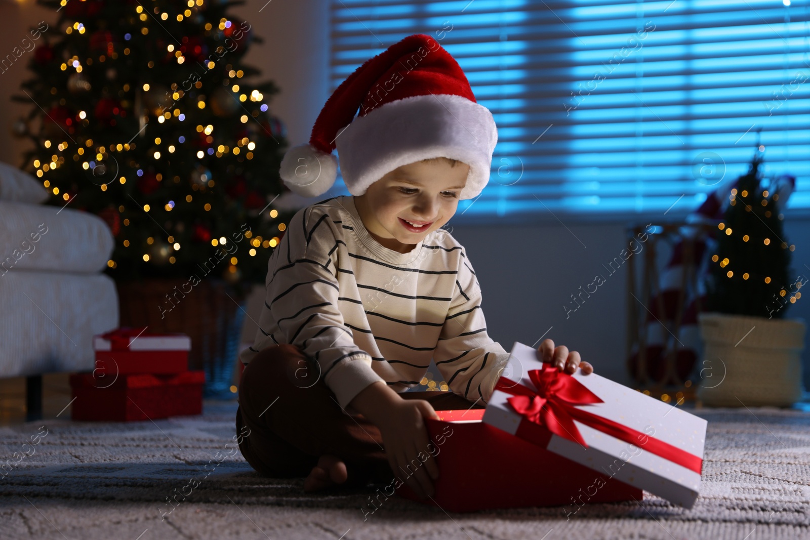 Photo of Happy little boy in Santa hat with Christmas gift on floor at home