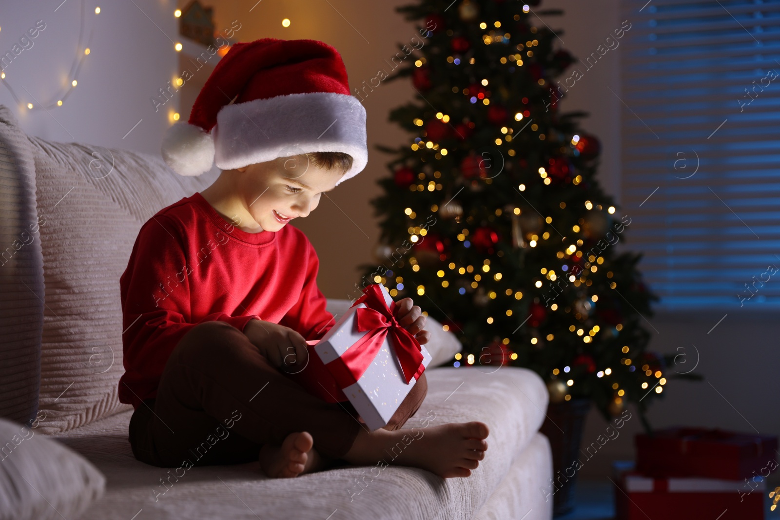 Photo of Happy little boy in Santa hat with Christmas gift on sofa at home. Space for text