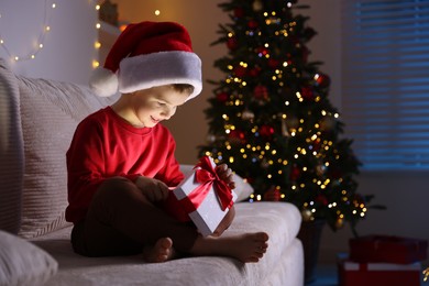 Photo of Happy little boy in Santa hat with Christmas gift on sofa at home. Space for text