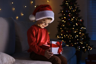 Photo of Happy little boy in Santa hat with Christmas gift on sofa at home. Space for text