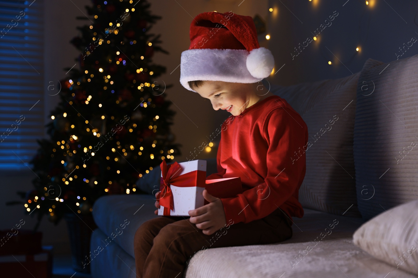 Photo of Happy little boy in Santa hat with Christmas gift on sofa at home. Space for text