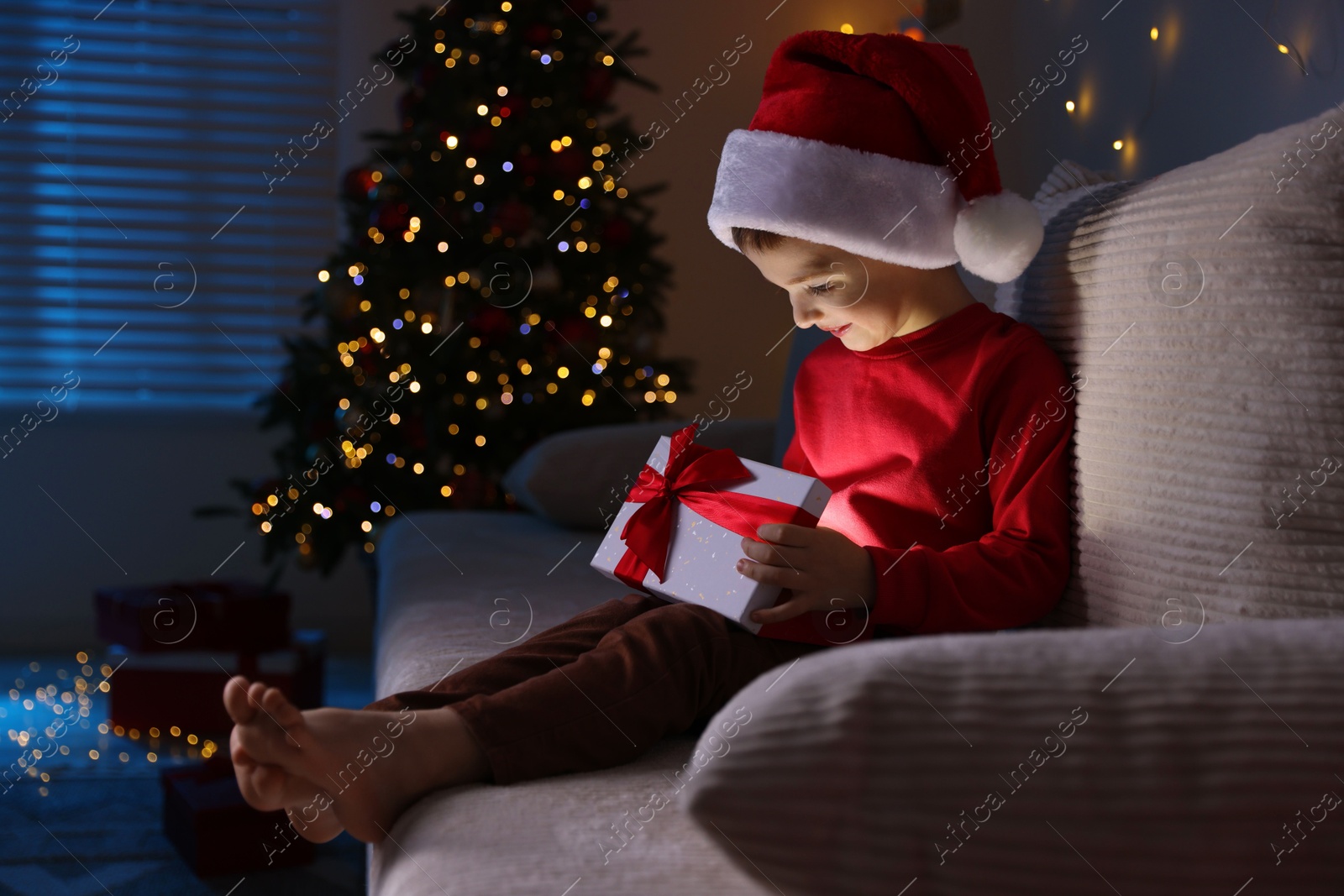 Photo of Happy little boy in Santa hat with Christmas gift on sofa at home