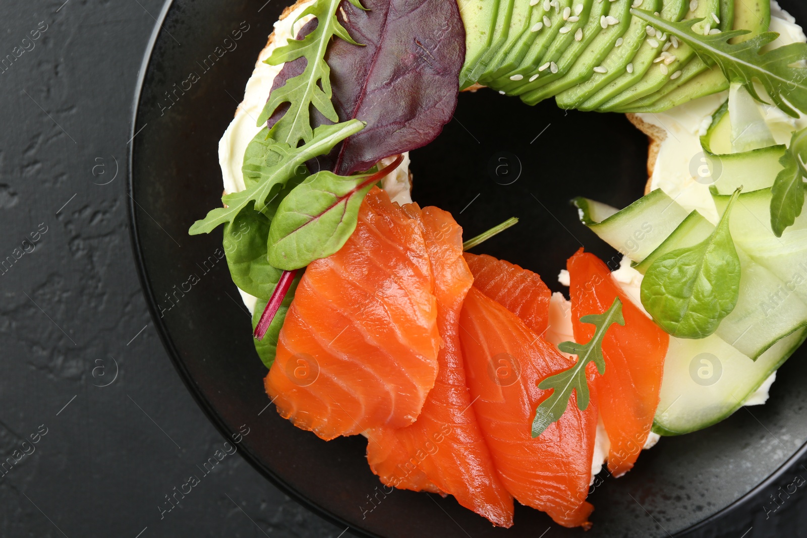 Photo of Delicious bagel with salmon, cream cheese, cucumber and avocado on black table, top view