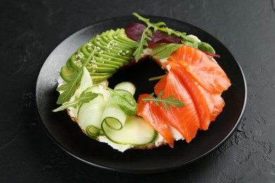 Photo of Delicious bagel with salmon, cream cheese, cucumber and avocado on black table, closeup