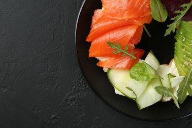 Photo of Delicious bagel with salmon, cream cheese, cucumber and avocado on black table, top view. Space for text