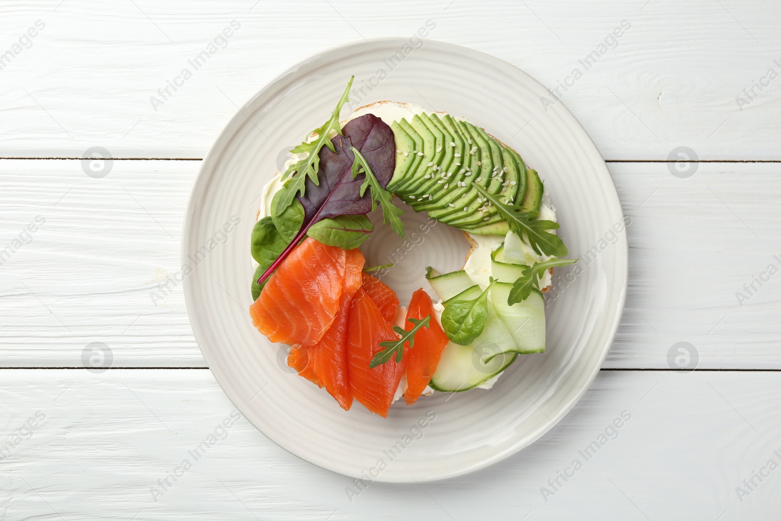 Photo of Delicious bagel with salmon, cream cheese, cucumber and avocado on white wooden table, top view