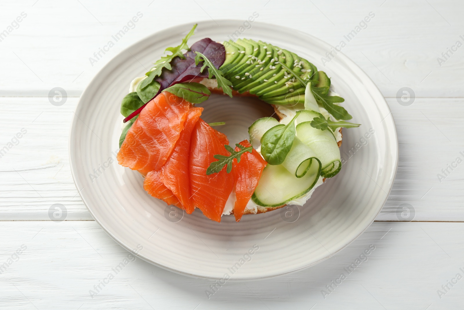 Photo of Delicious bagel with salmon, cream cheese, cucumber and avocado on white wooden table, closeup