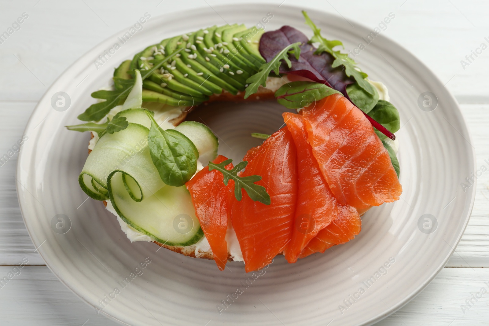 Photo of Delicious bagel with salmon, cream cheese, cucumber and avocado on white wooden table, closeup