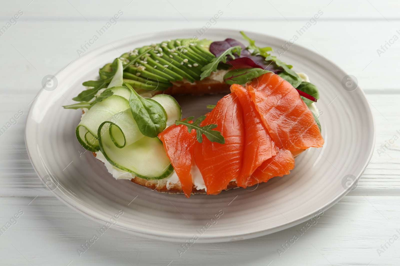 Photo of Delicious bagel with salmon, cream cheese, cucumber and avocado on white wooden table, closeup