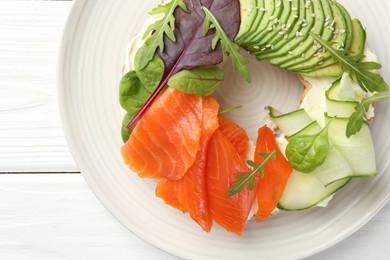 Photo of Delicious bagel with salmon, cream cheese, cucumber and avocado on white wooden table, top view