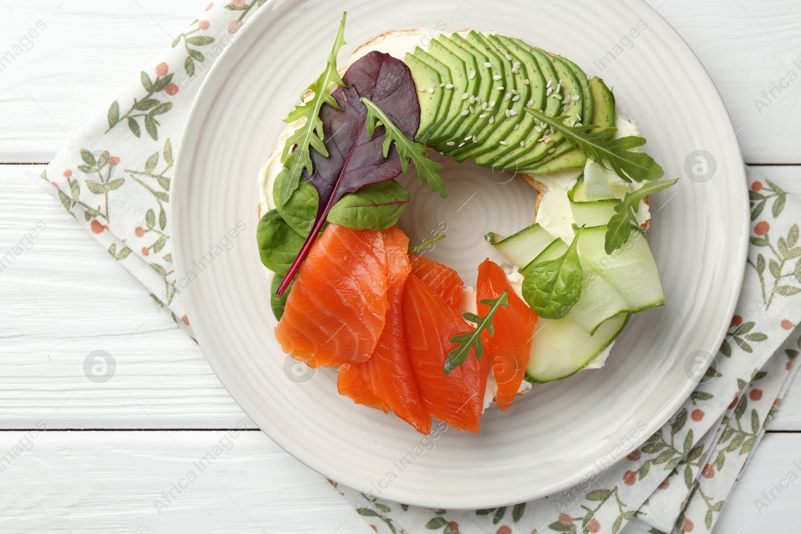 Photo of Delicious bagel with salmon, cream cheese, cucumber and avocado on white wooden table, top view