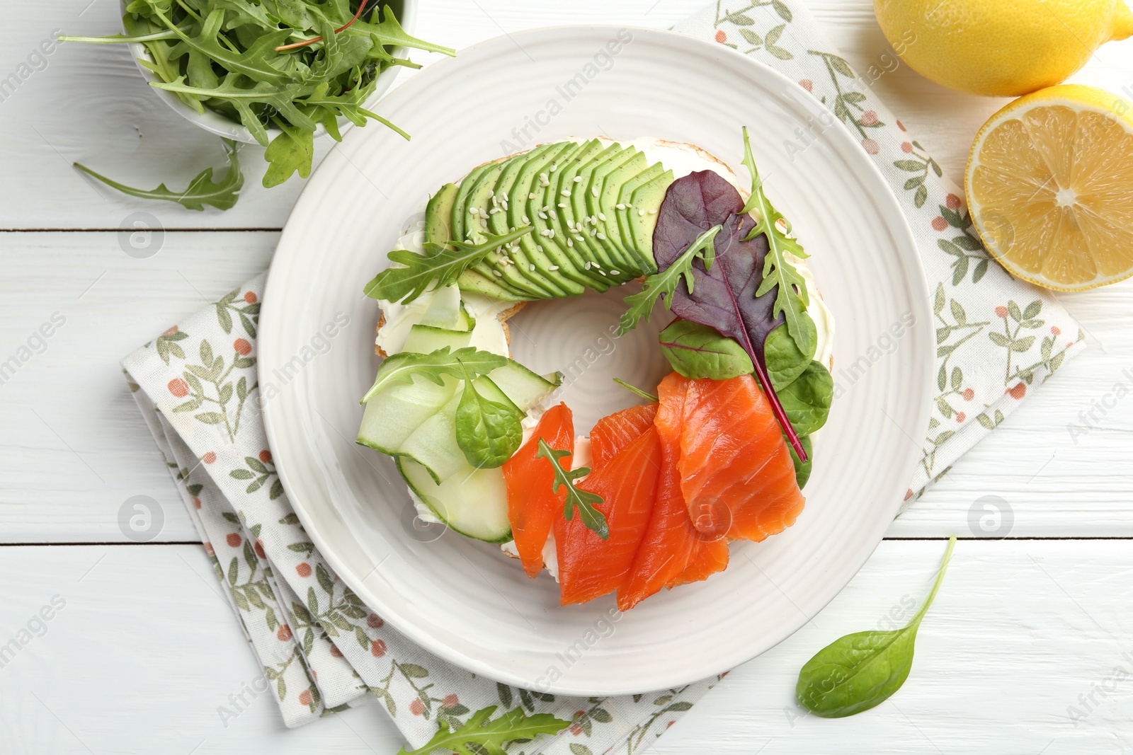 Photo of Delicious bagel with salmon, cream cheese, cucumber and avocado on white wooden table, flat lay