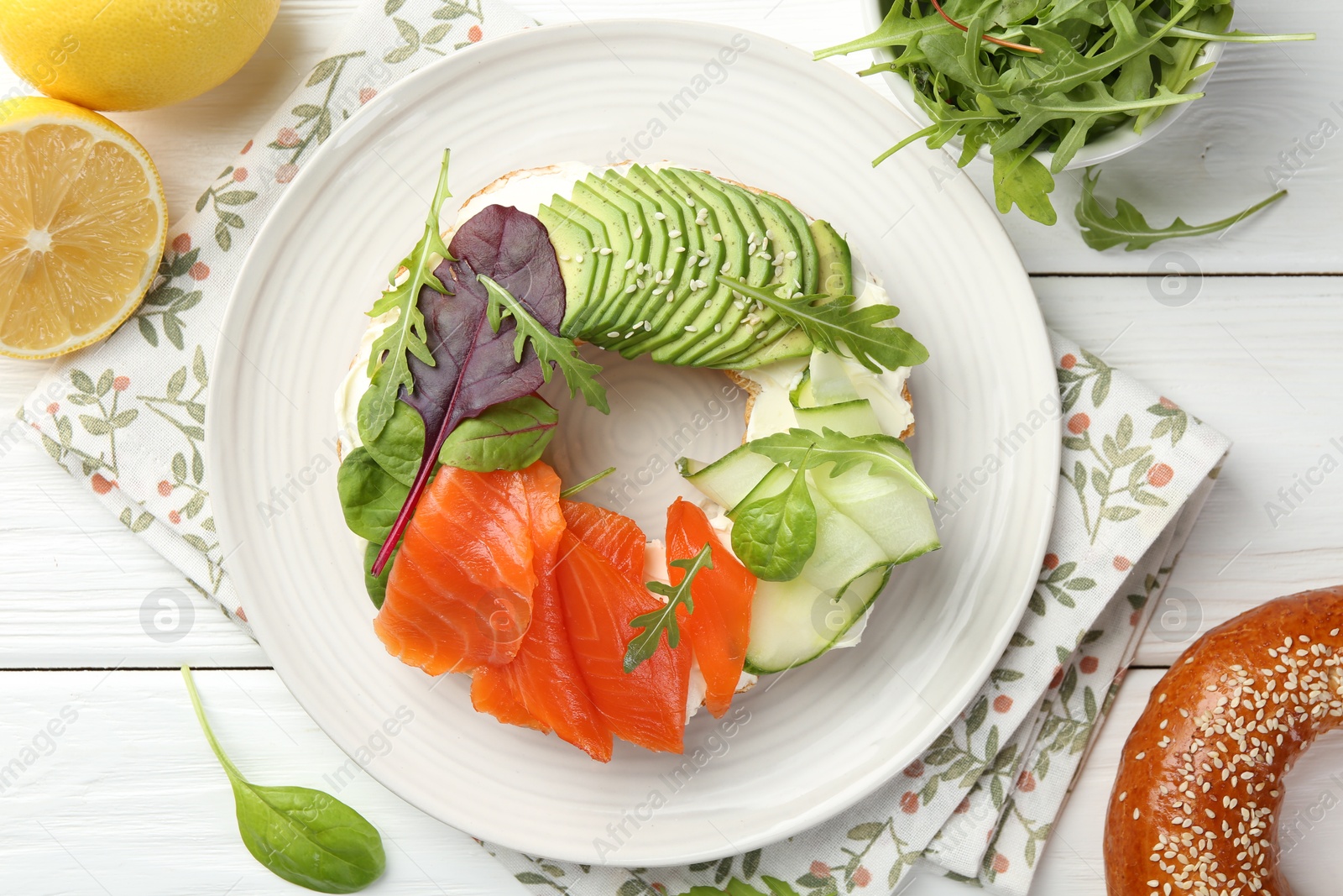 Photo of Delicious bagel with salmon, cream cheese, cucumber and avocado on white wooden table, flat lay