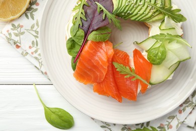 Photo of Delicious bagel with salmon, cream cheese, cucumber and avocado on white wooden table, flat lay