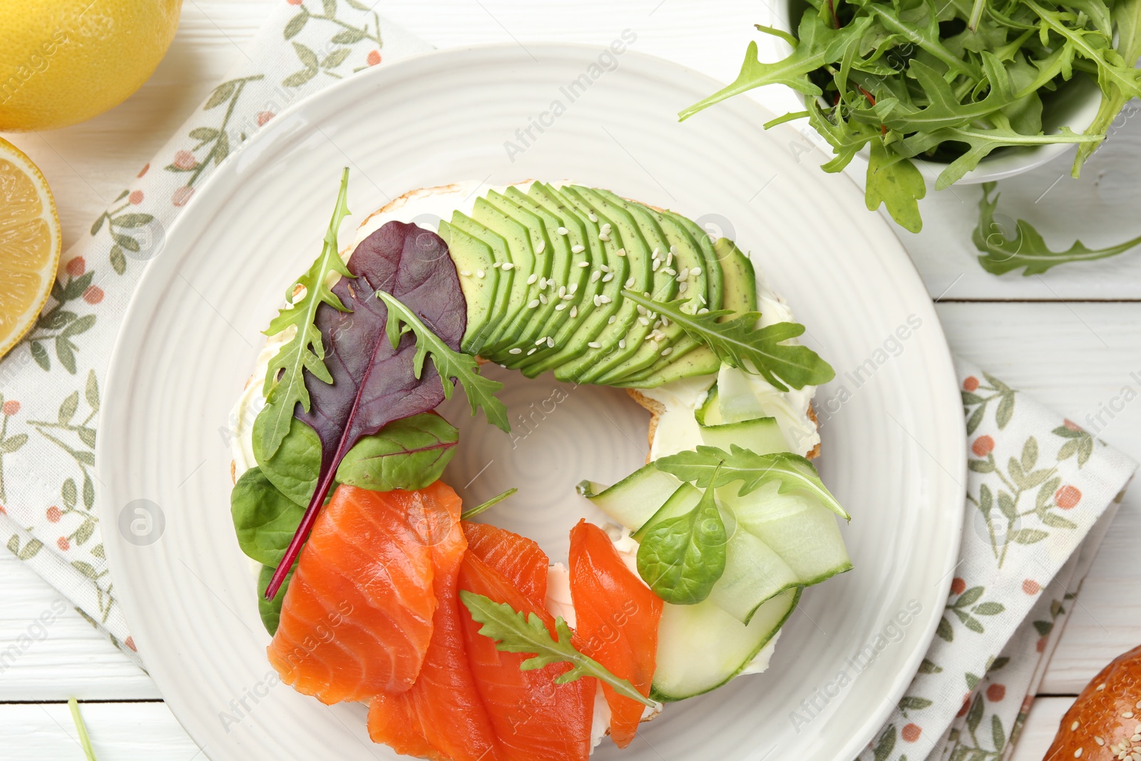 Photo of Delicious bagel with salmon, cream cheese, cucumber and avocado on white wooden table, flat lay