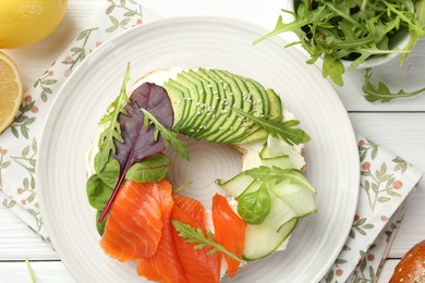 Photo of Delicious bagel with salmon, cream cheese, cucumber and avocado on white wooden table, flat lay