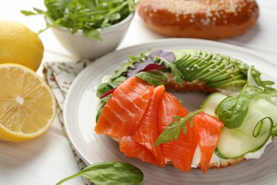 Photo of Delicious bagel with salmon, cream cheese, cucumber and avocado on white wooden table, closeup