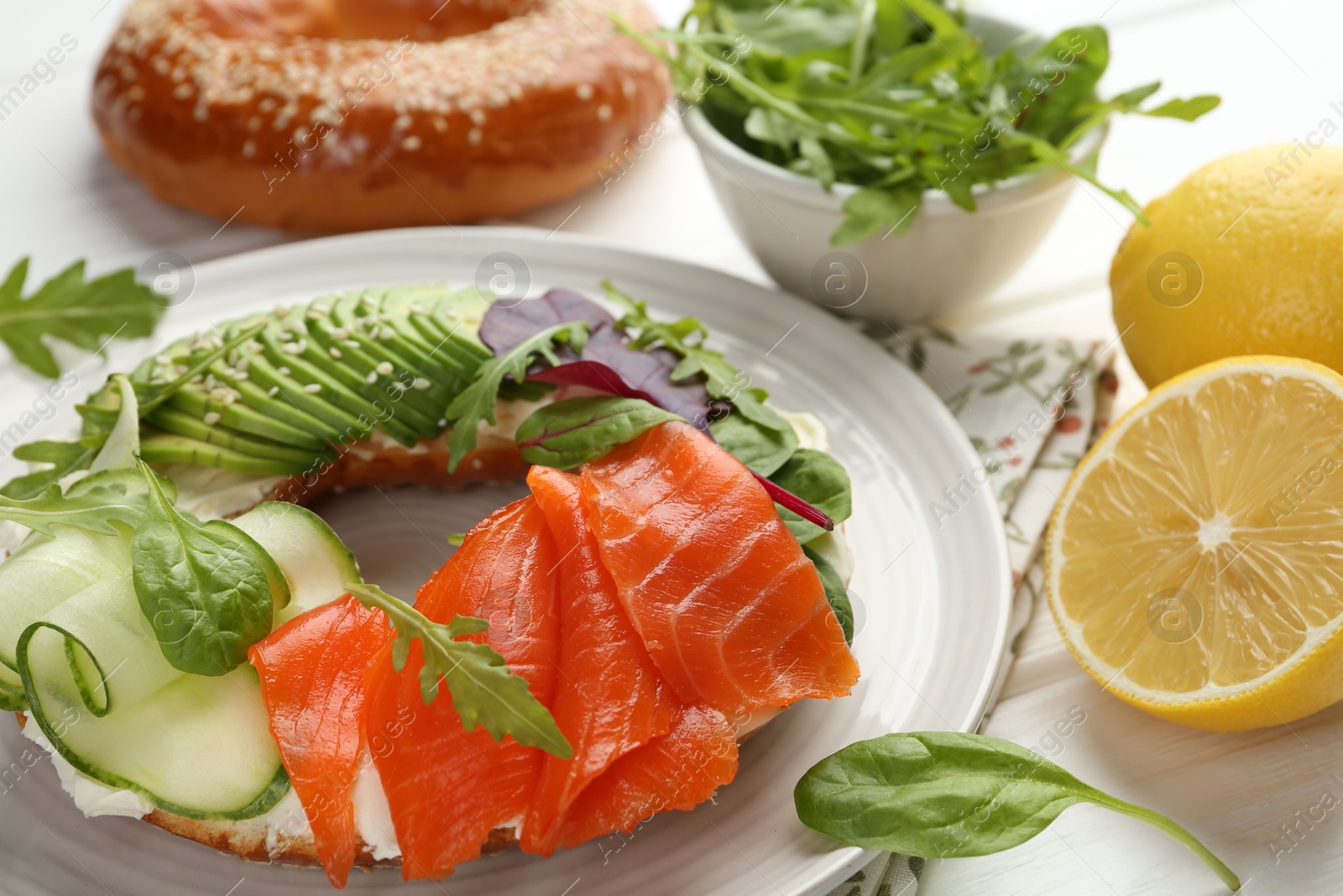 Photo of Delicious bagel with salmon, cream cheese, cucumber and avocado on white wooden table, closeup