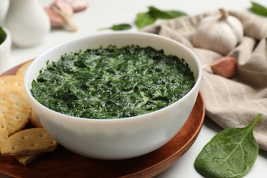 Delicious spinach sauce in bowl, crackers and garlic on white table, closeup