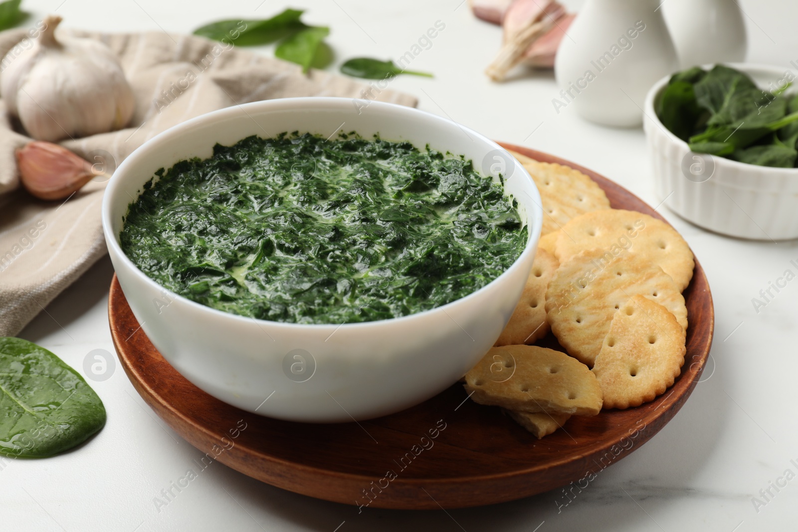 Photo of Delicious spinach sauce in bowl, crackers and garlic on white marble table, closeup