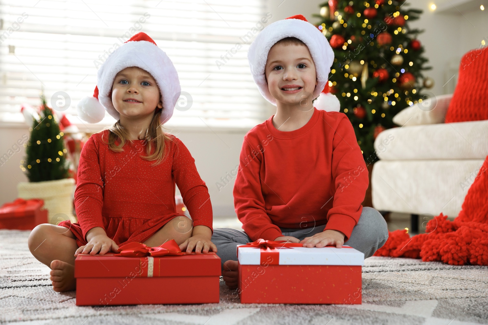 Photo of Happy little children in Santa hats with Christmas gifts on floor at home