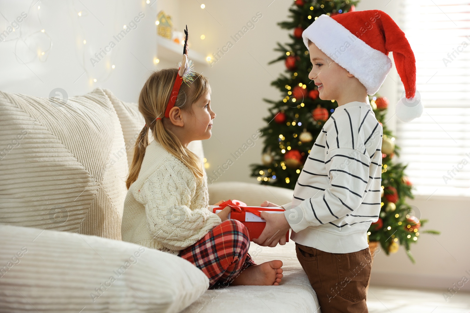 Photo of Happy little boy in Santa hat presenting his sister with Christmas gift at home