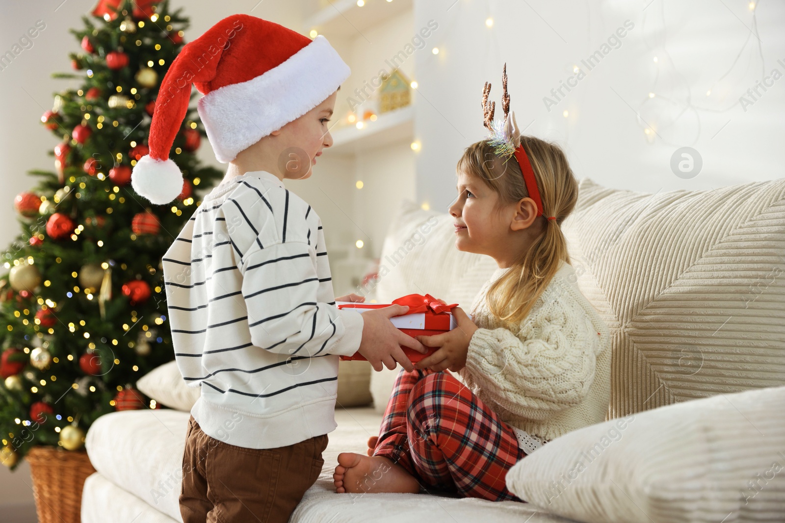 Photo of Happy little boy in Santa hat presenting his sister with Christmas gift at home