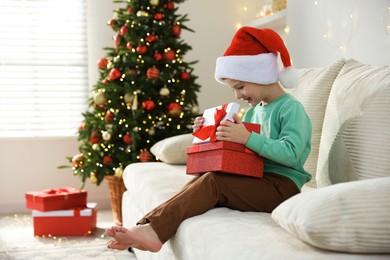 Photo of Happy little boy in Santa hat with Christmas gifts on sofa at home