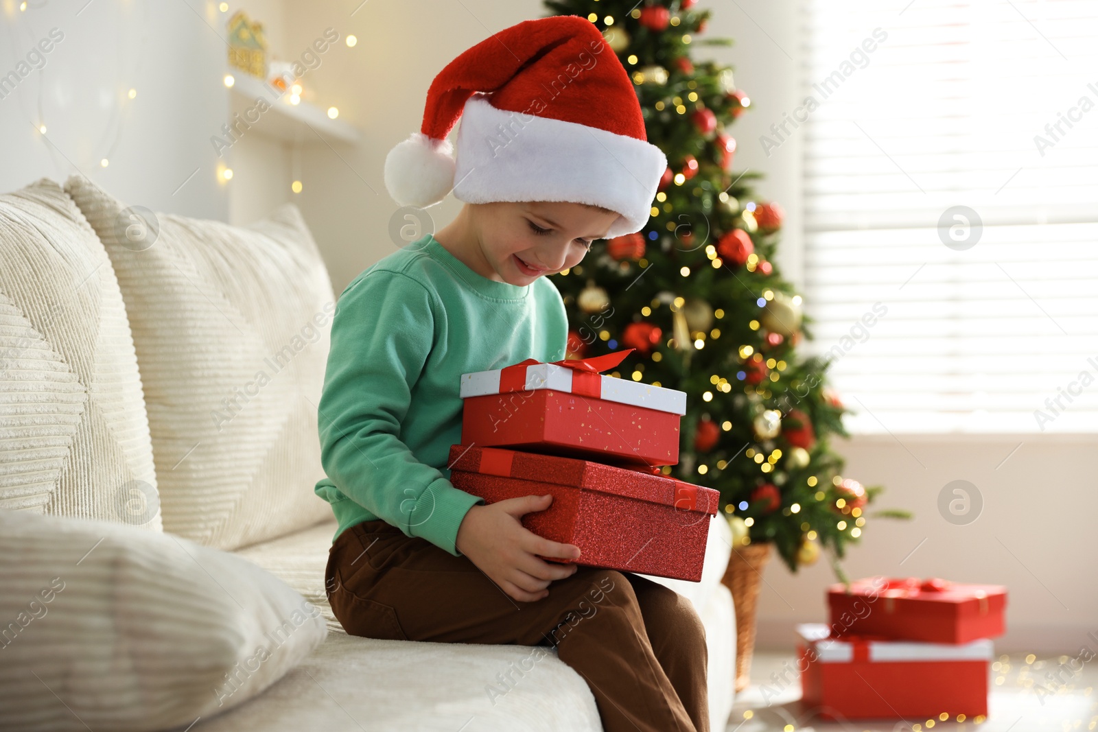 Photo of Happy little boy in Santa hat with Christmas gifts on sofa at home