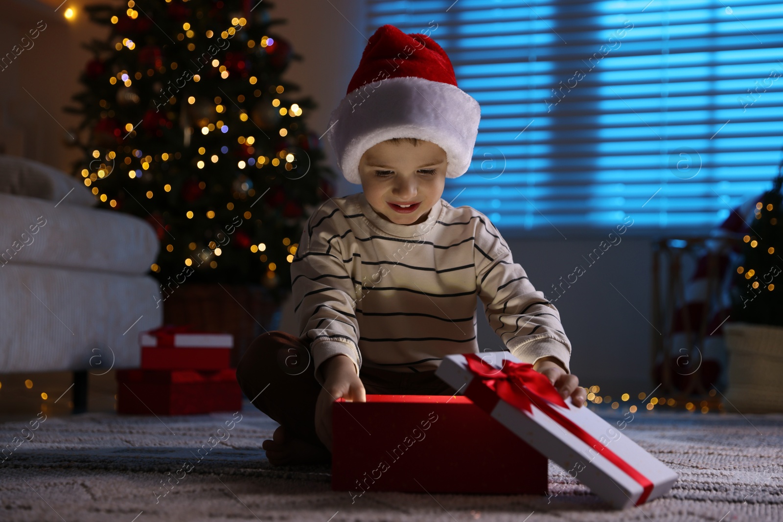 Photo of Happy little boy in Santa hat with Christmas gift on floor at home