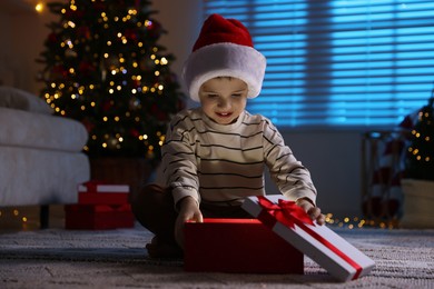 Photo of Happy little boy in Santa hat with Christmas gift on floor at home