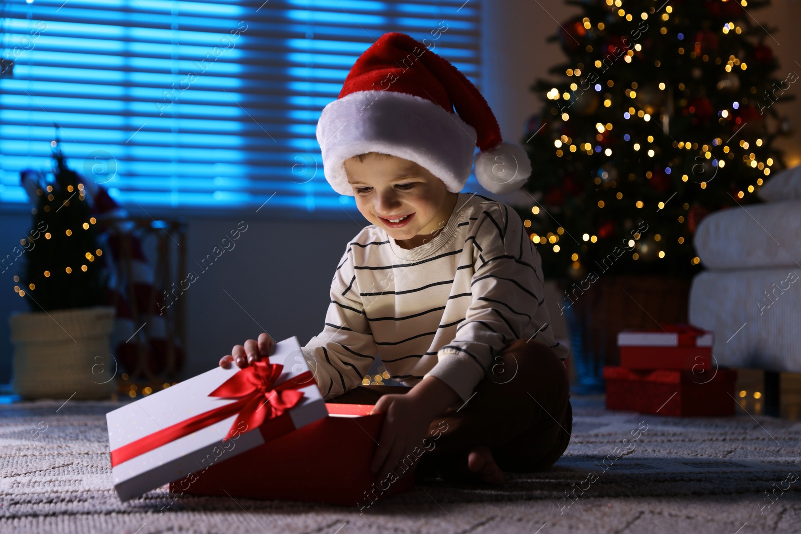 Photo of Happy little boy in Santa hat with Christmas gift on floor at home