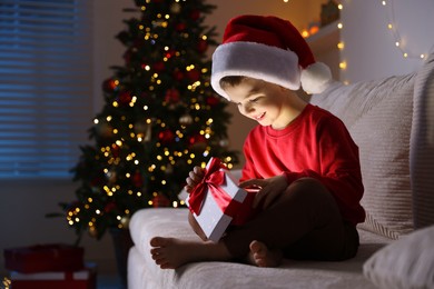 Photo of Happy little boy in Santa hat with Christmas gift on sofa at home. Space for text