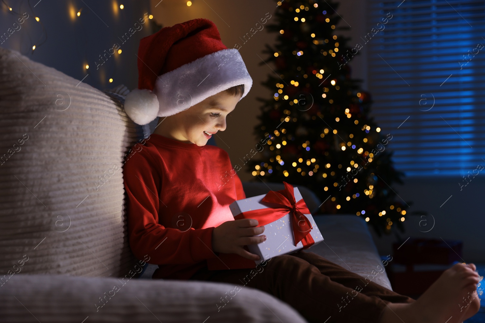 Photo of Happy little boy in Santa hat with Christmas gift on sofa at home