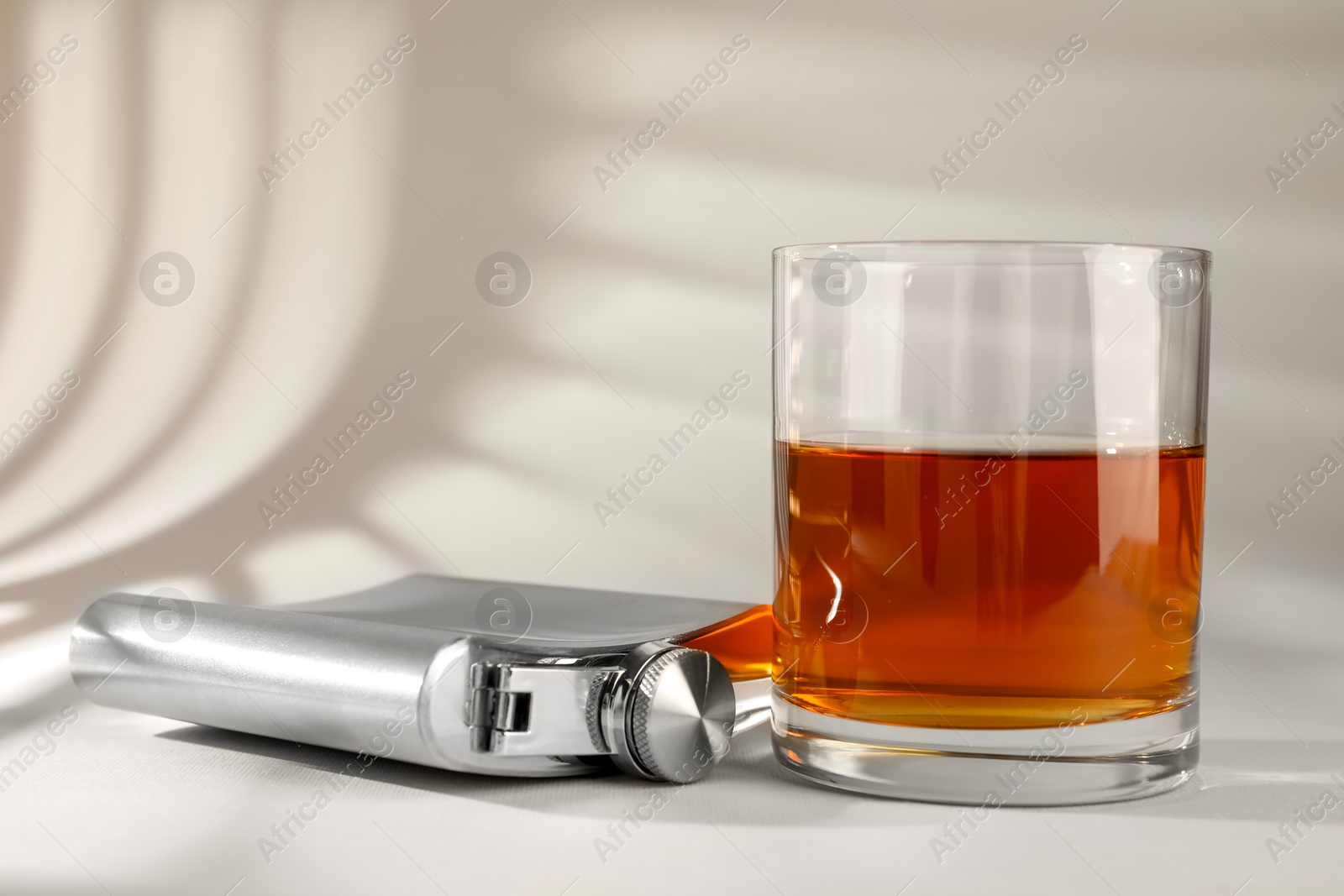 Photo of Hip flask and glass of whiskey on light table, closeup