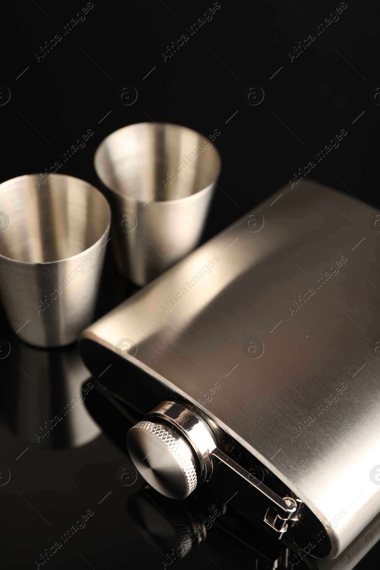 Photo of Hip flask and cups on black mirror surface, closeup