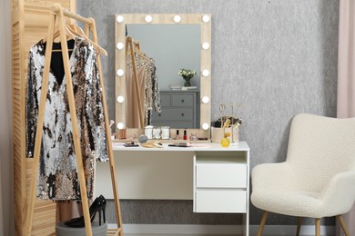 Photo of Makeup room. Mirror, dressing table, different beauty products, clothing rack and armchair indoors