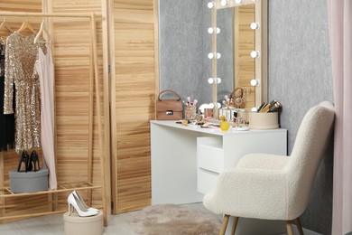 Photo of Makeup room. Mirror, dressing table, different beauty products, clothing rack and armchair indoors