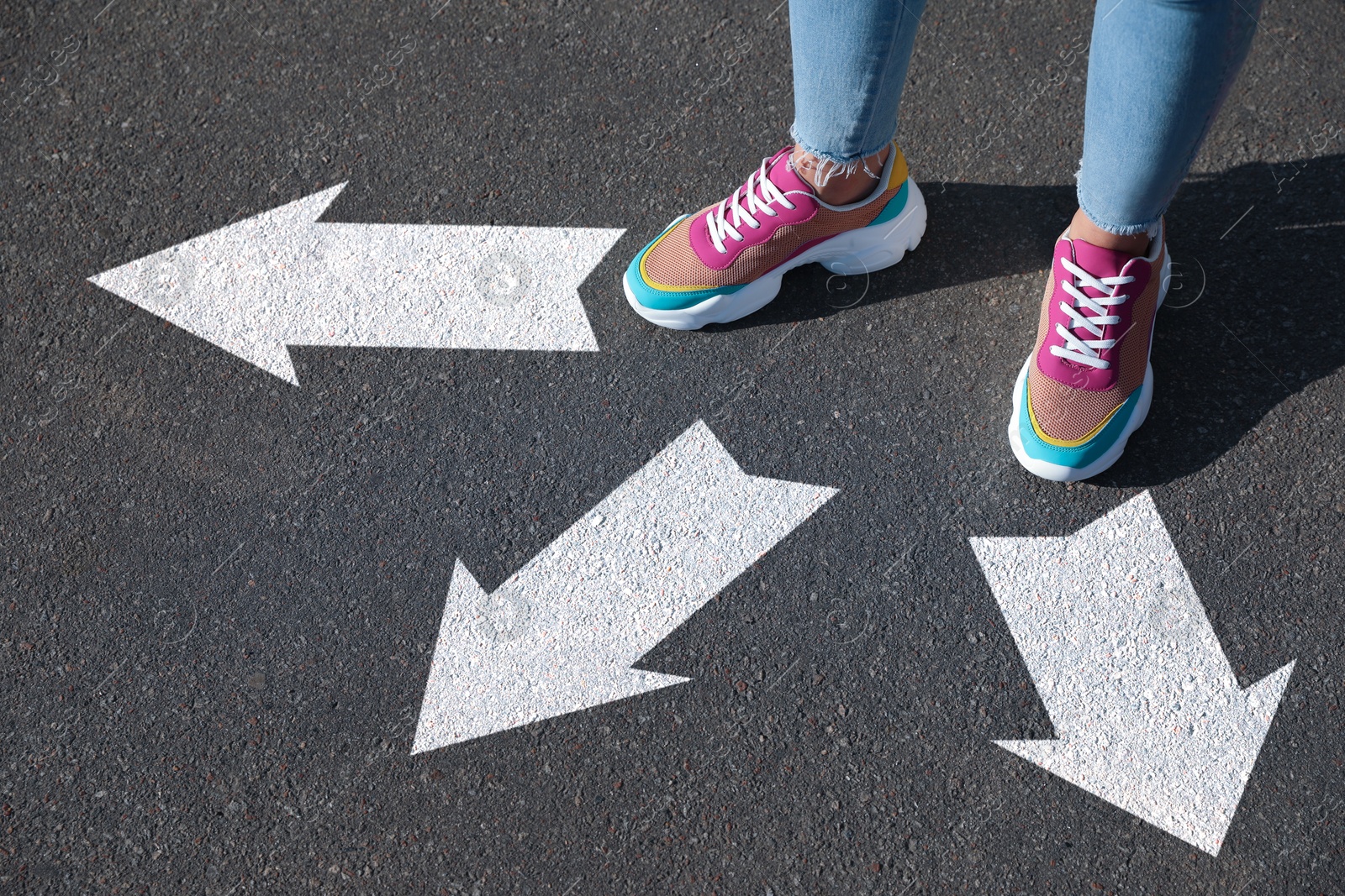 Image of Woman standing near white arrows pointing in different directions on asphalt road, closeup. Concept of choice and making decisions