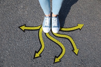 Image of Woman standing on asphalt road with yellow arrows pointing in different directions, top view. Concept of choice and making decisions