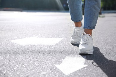 Image of Woman walking toward one of two white arrows pointing in different directions on asphalt road, closeup. Concept of choice and making decisions