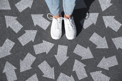 Image of Woman standing on asphalt road with white arrows pointing in different directions, top view. Concept of choice and making decisions