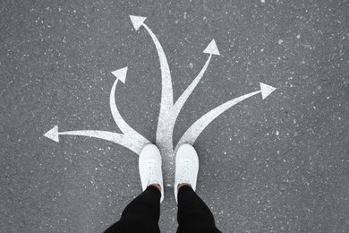 Image of Woman standing on asphalt road with white arrows pointing in different directions, top view. Concept of choice and making decisions