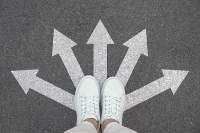 Image of Woman standing on asphalt road with white arrows pointing in different directions, top view. Concept of choice and making decisions