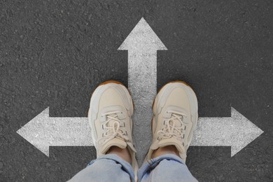 Image of Woman standing on asphalt road with white arrows pointing in different directions, top view. Concept of choice and making decisions