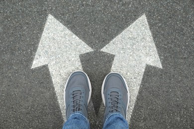 Image of Man standing on asphalt road with white arrows pointing in different directions, top view. Concept of choice and making decisions