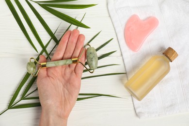 Photo of Woman with face roller, gua sha tool, cosmetic product, towel and leaves on white wooden background, top view