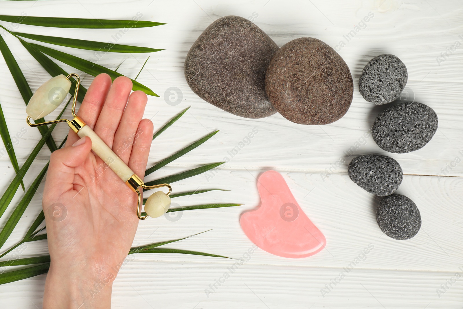 Photo of Woman with face roller, gua sha tool, stones and leaves on white wooden background, top view